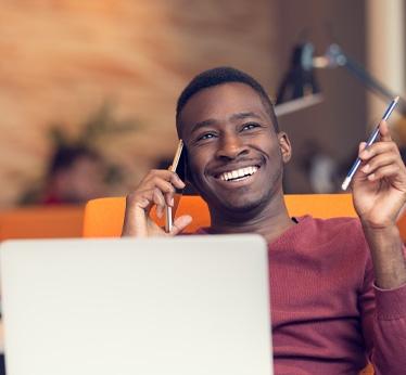 Man smiling while talking on his smartphone and sitting at his desk with his laptop